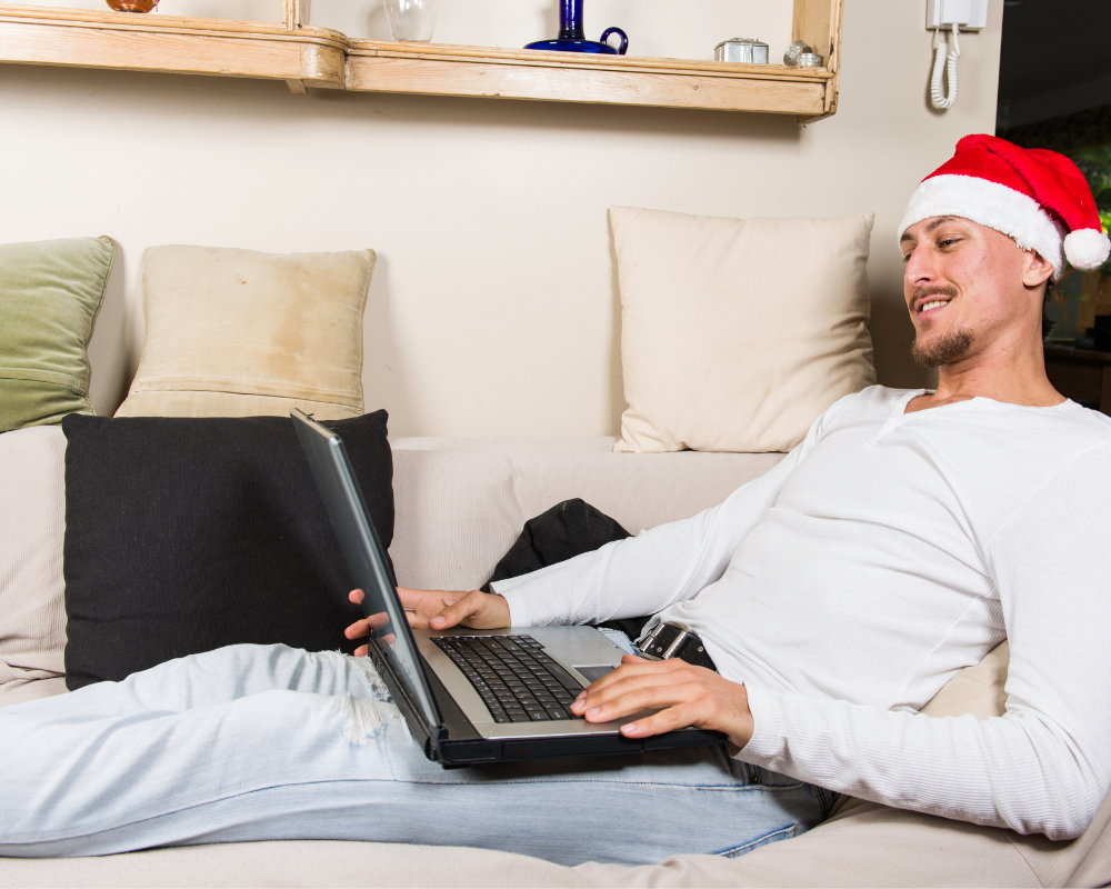 Man sitting on the sofa with a laptop wearing a Christmas hat.