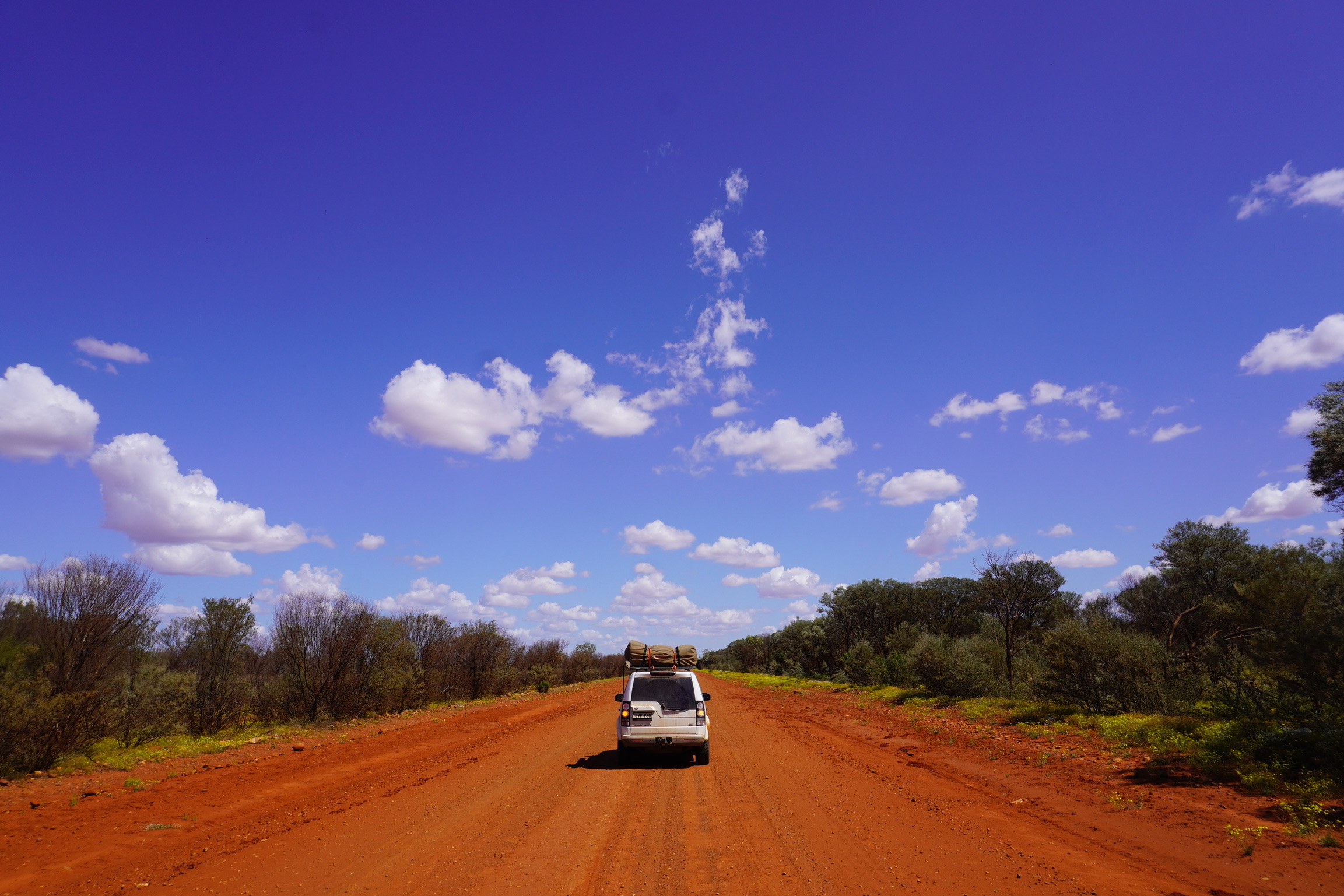 A 4WD packed for adventure drives down a red dirt road in the Australian outback under a bright blue sky with scattered clouds