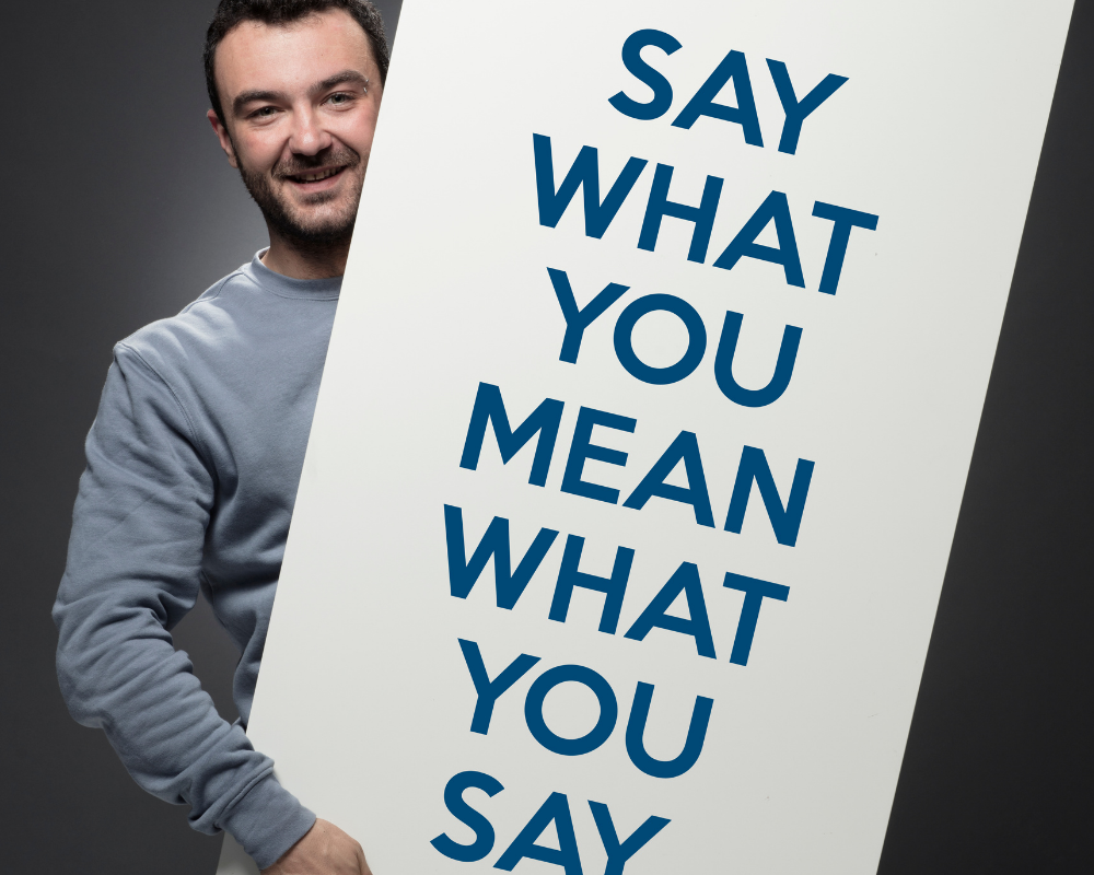 Man in blue shirt holding sign with words say what you mean what you say