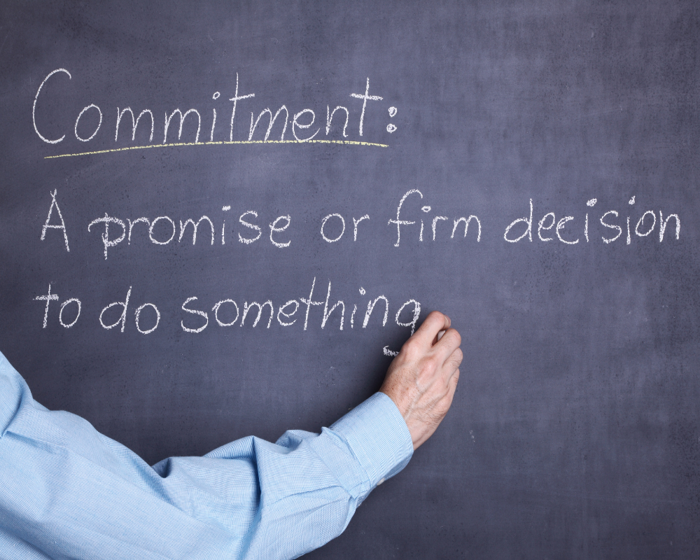 Man in blue shirt writing the meaning of commitment on a chalk board.