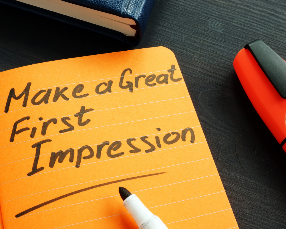 An orange notepad with “Make a Great First Impression” handwritten in black marker, symbolizing the importance of professional branding in job searching and hiring. A whiteboard marker and an orange highlighter sit on a dark wooden desk.